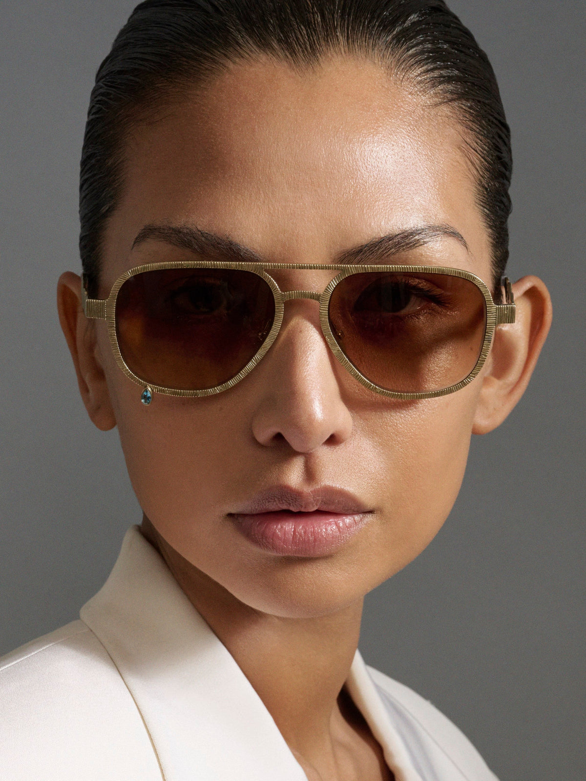 A close-up portrait of a model wearing aviator-style sunglasses with a textured gold frame and dark brown lenses, paired with a white blazer against a neutral gray background.