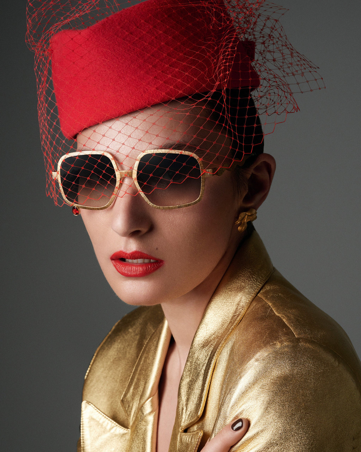 A close-up portrait of a model wearing a bright red felt fascinator hat with a red net veil, gold-framed aviator-style sunglasses, red lipstick, and a shiny gold jacket against a dark gray studio background.