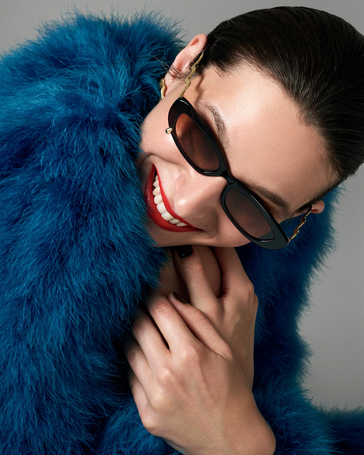 A close-up lifestyle shot of a model wearing black cat-eye sunglasses with gold chain accents and a vibrant blue faux fur coat against a solid gray background.
