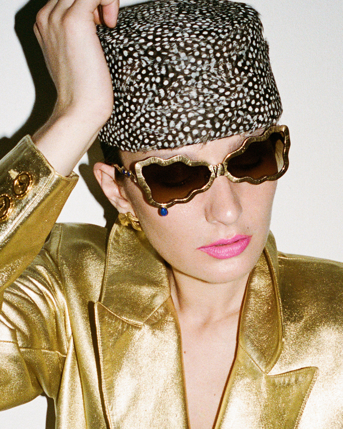 A close-up fashion shot of a model wearing a metallic gold satin blazer, a matching gold hat with white spots, and scalloped gold sunglasses against a plain light background.