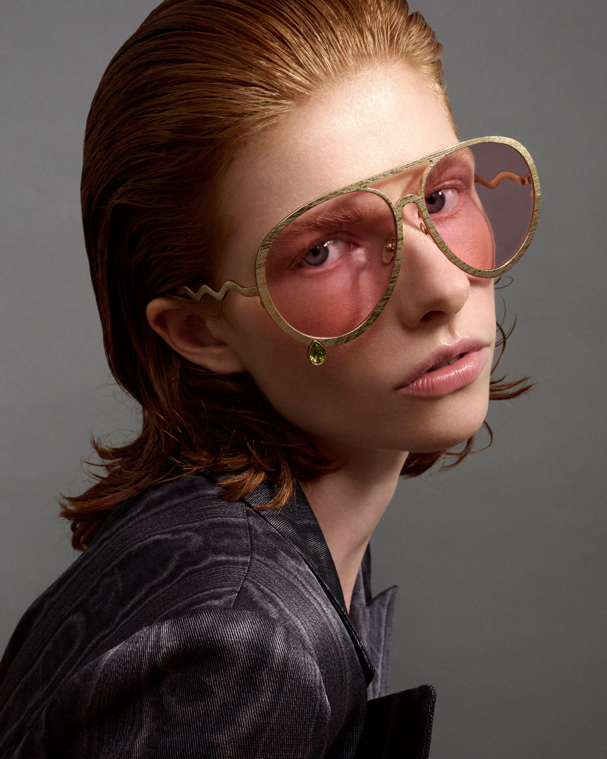 A close-up portrait of a model with reddish-brown hair wearing oversized aviator-style sunglasses with wavy gold metal frames and pink-tinted lenses, against a dark gray studio background.