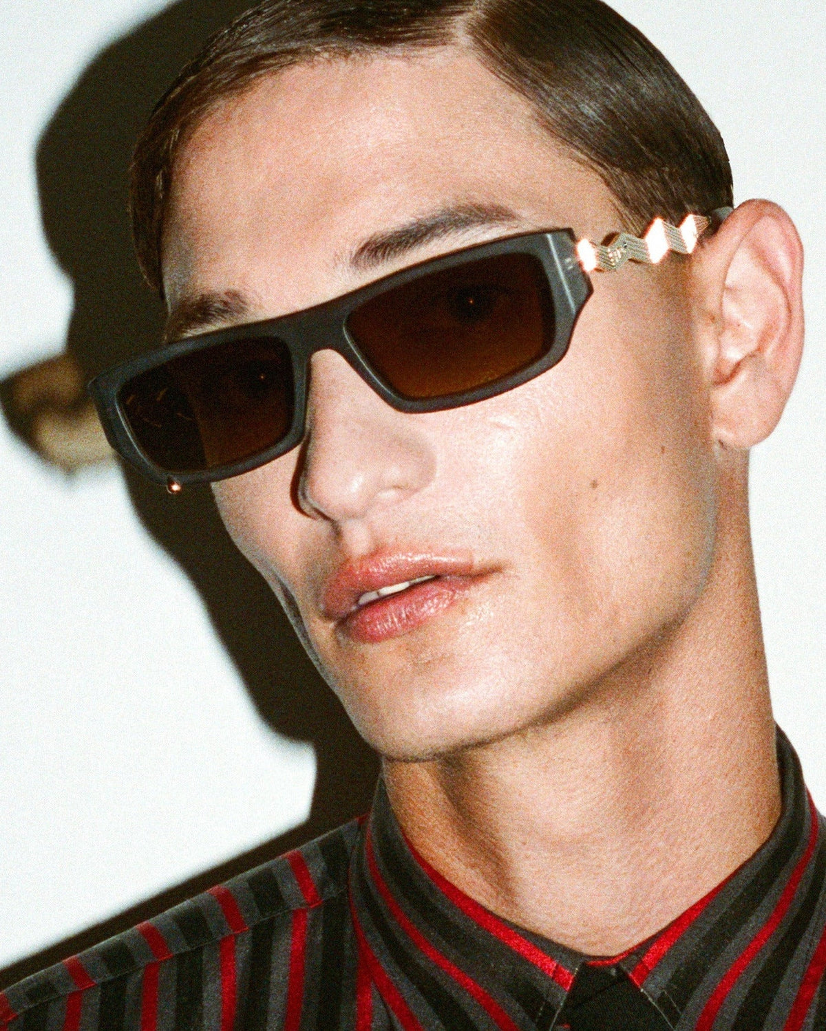A close-up portrait of a man wearing dark rectangular sunglasses with a unique gold-toned temple detail and a black-and-red vertically striped shirt with a black tie, set against a plain off-white background.