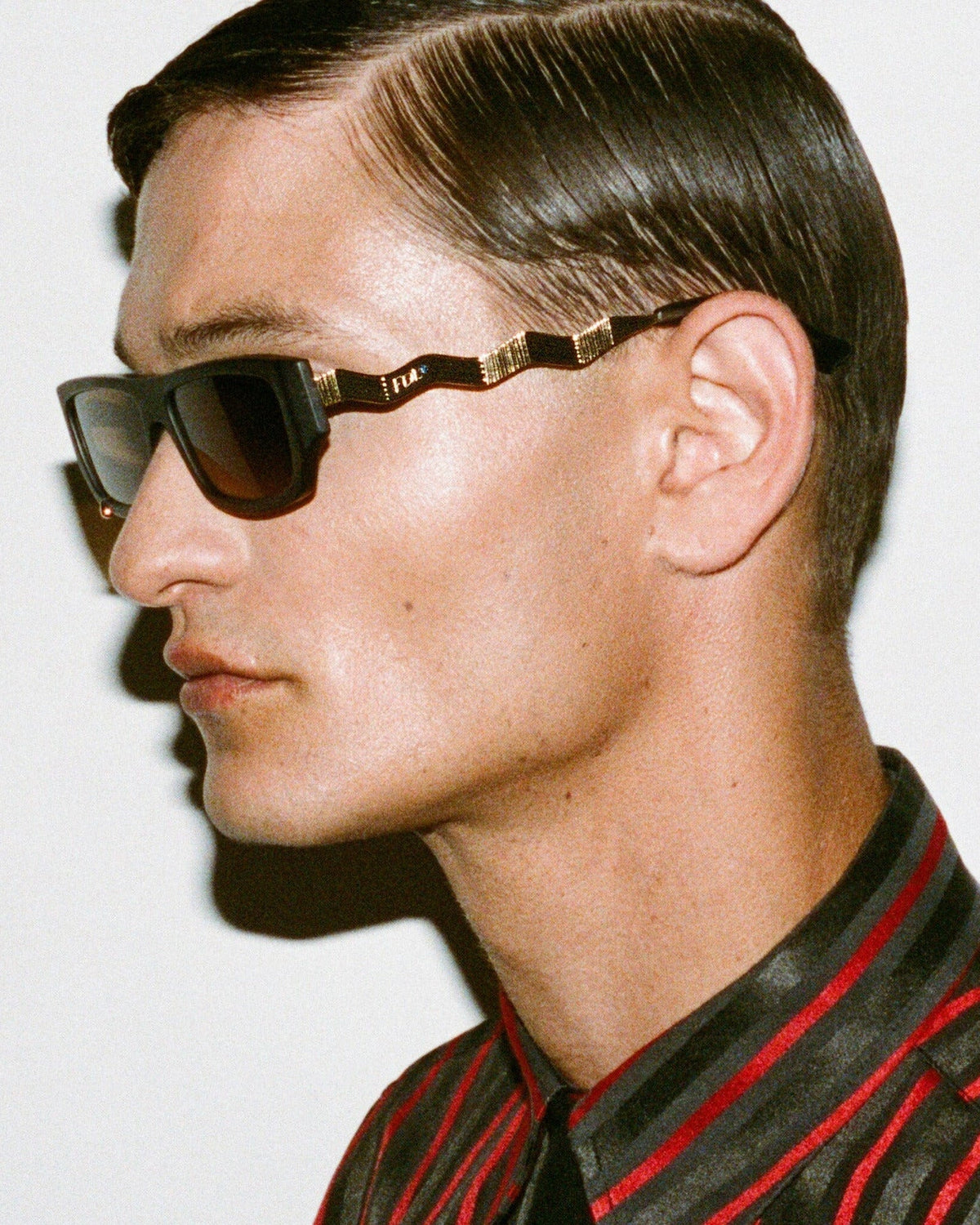 A man in profile wearing dark sunglasses with gold-tone metal temples and a red-and-black striped shirt against a plain light background.