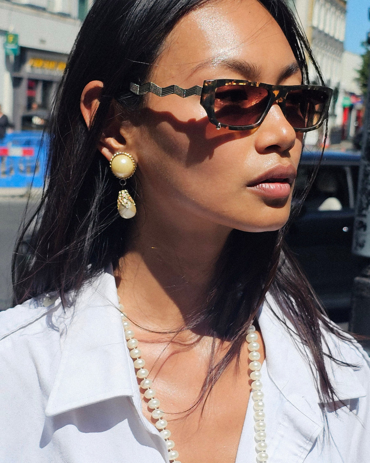 A close-up lifestyle shot of a woman wearing tortoiseshell sunglasses with gold geometric metal frames, gold drop earrings, and a pearl necklace, set against a blurred city street background.