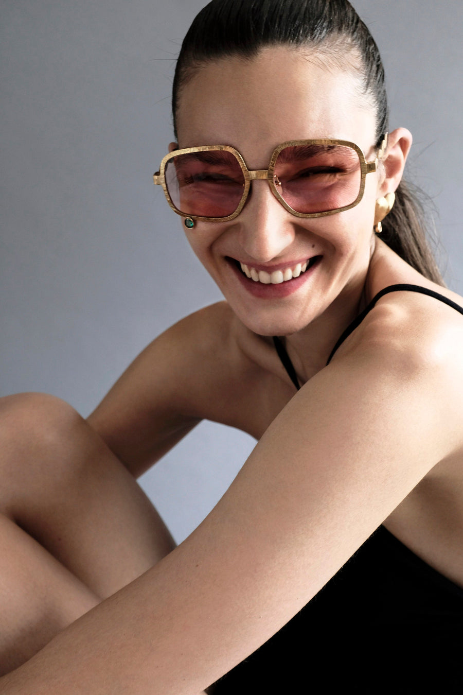 A smiling woman wearing oversized square sunglasses with gold frames and pink-tinted lenses, posed against a plain gray background.