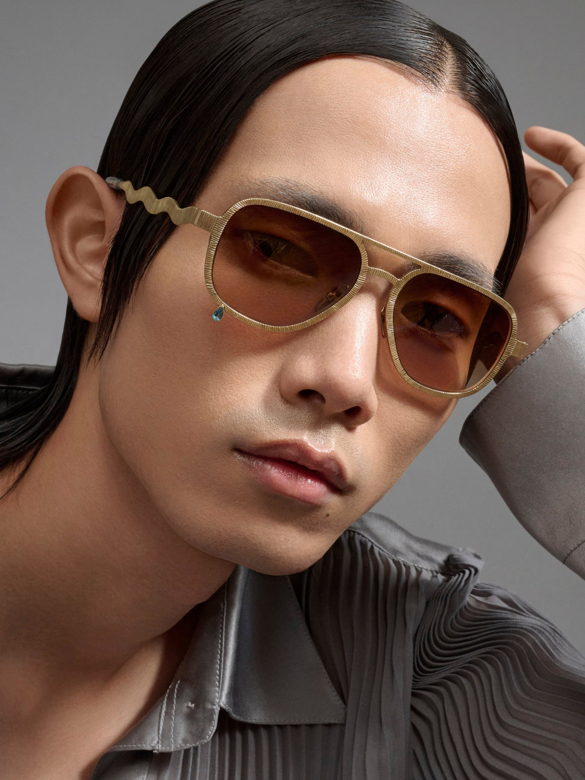 A close-up portrait of a man wearing stylish gold-framed sunglasses with brown lenses and a small turquoise accent, paired with a gray striped shirt against a neutral gray background.