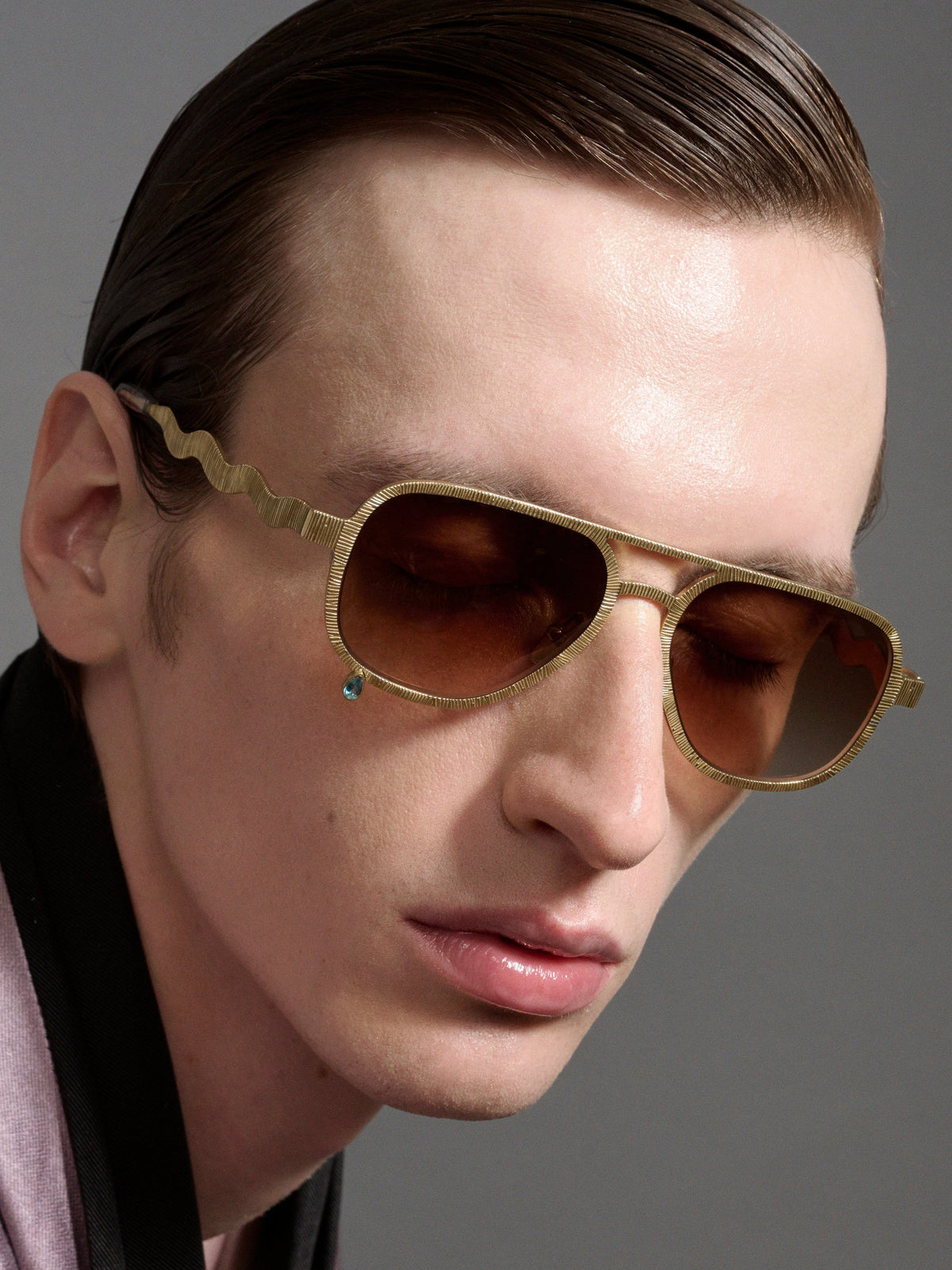 A close-up of a man wearing Gucci aviator sunglasses with a gold-tone wavy metal frame and brown lenses, set against a solid gray background.
