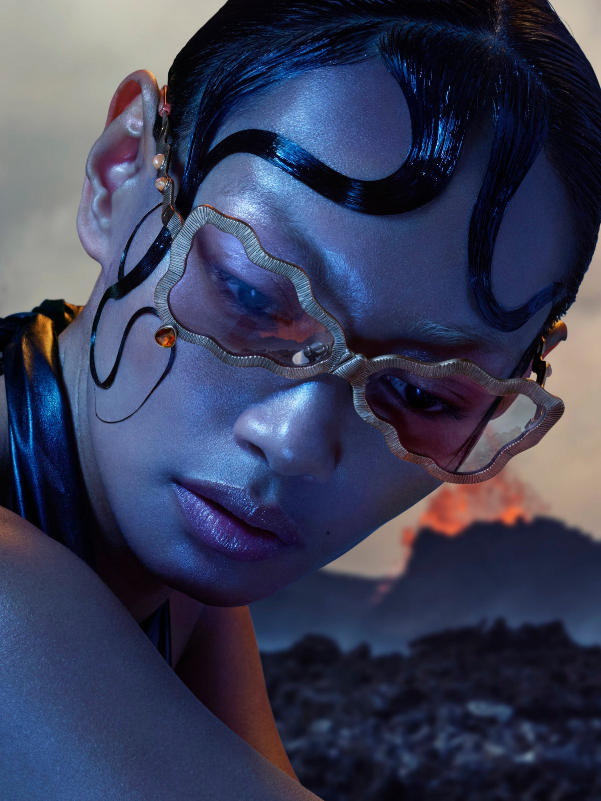 A close-up portrait of a model wearing distinctive gold-framed geometric eyeglasses with amber-tinted lenses, styled with dramatic dark hair and face makeup against a moody, volcanic outdoor background.