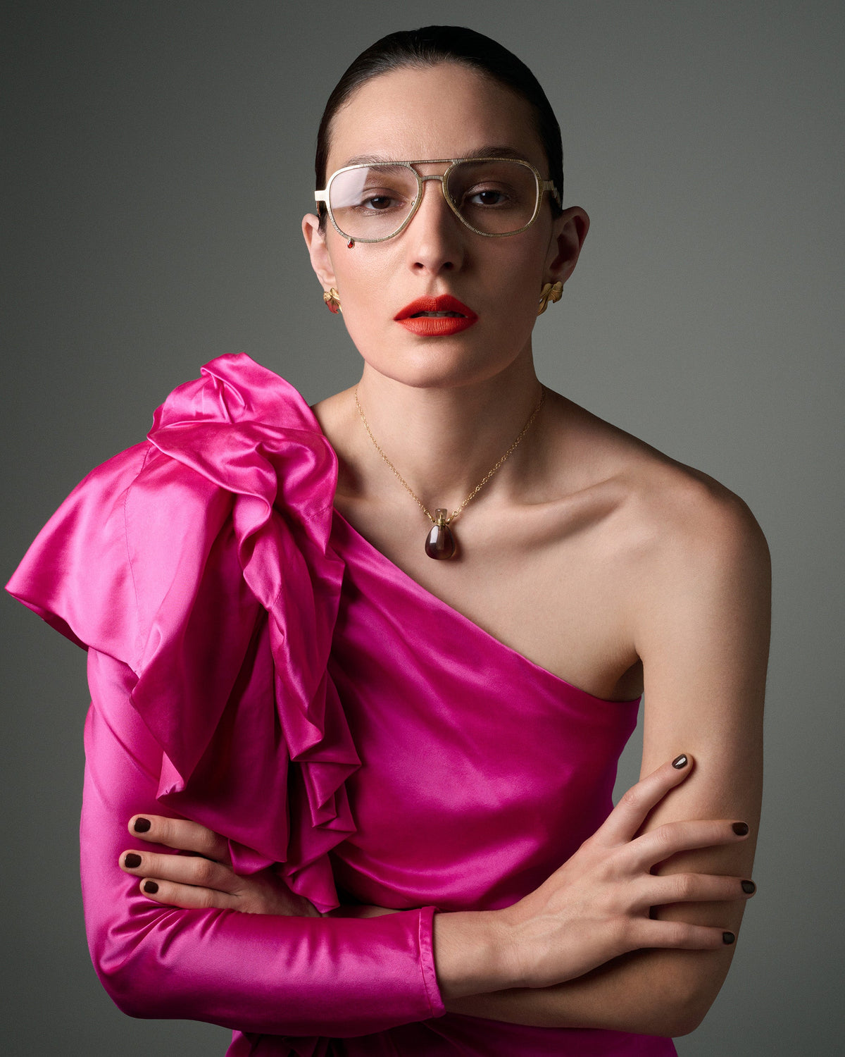 A woman with her hair pulled back wears clear-framed eyeglasses, a gold pendant necklace, and a bright fuchsia one-shoulder satin dress with a large ruffled bow on the shoulder. She is posed against a neutral gray studio background.