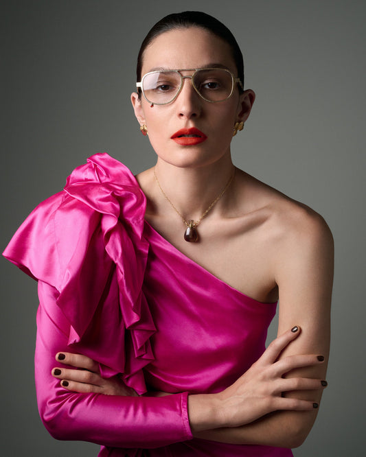 A woman with her hair pulled back wears clear-framed eyeglasses, a gold pendant necklace, and a bright fuchsia one-shoulder satin dress with a large ruffled bow on the shoulder. She is posed against a neutral gray studio background.
