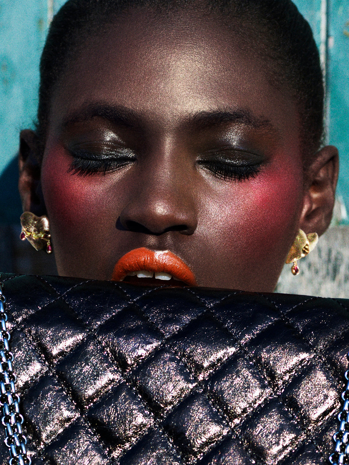 A close-up portrait of a model with dramatic eye makeup, pink blush, and bright orange lipstick, wearing gold earrings and holding a black quilted metallic clutch bag in front of their mouth.