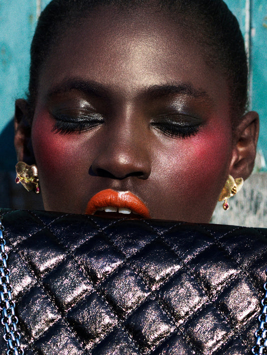 A close-up portrait of a model with dramatic eye makeup, pink blush, and bright orange lipstick, wearing gold earrings and holding a black quilted metallic clutch bag in front of their mouth.