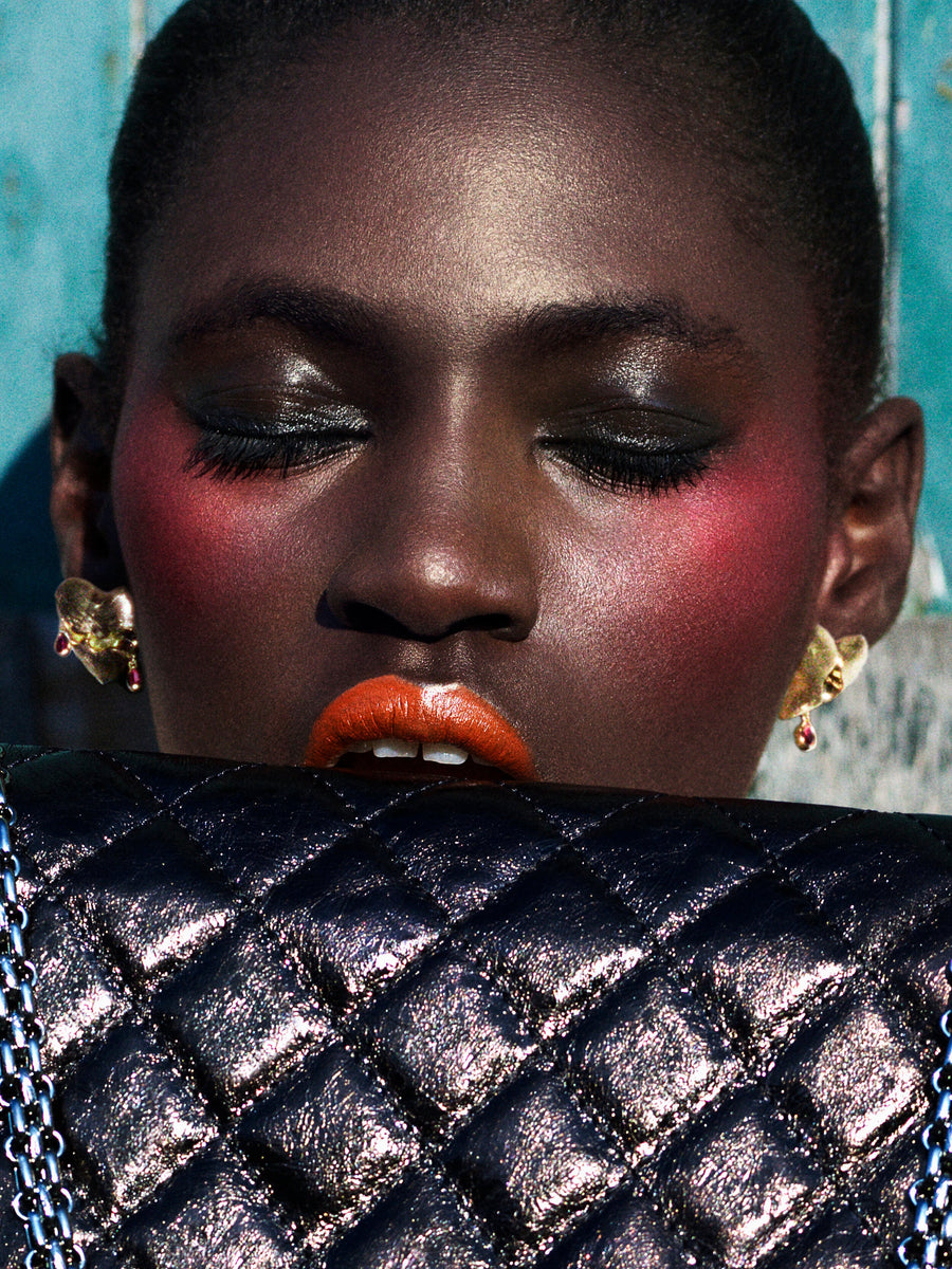 A close-up portrait of a model with dramatic eye makeup, pink blush, and bright orange lipstick, wearing gold earrings and holding a black quilted metallic clutch bag in front of their mouth.