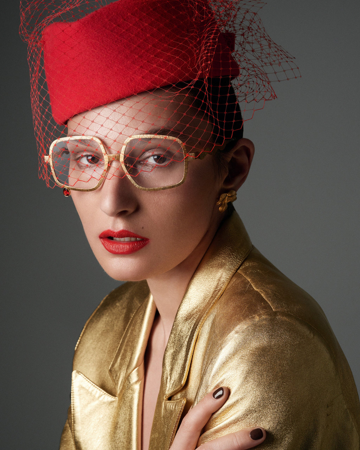 A model wearing a metallic gold satin jacket, gold-rimmed square aviator glasses, a red felt fascinator hat with a red net veil, and red lipstick, posed against a dark gray studio background.