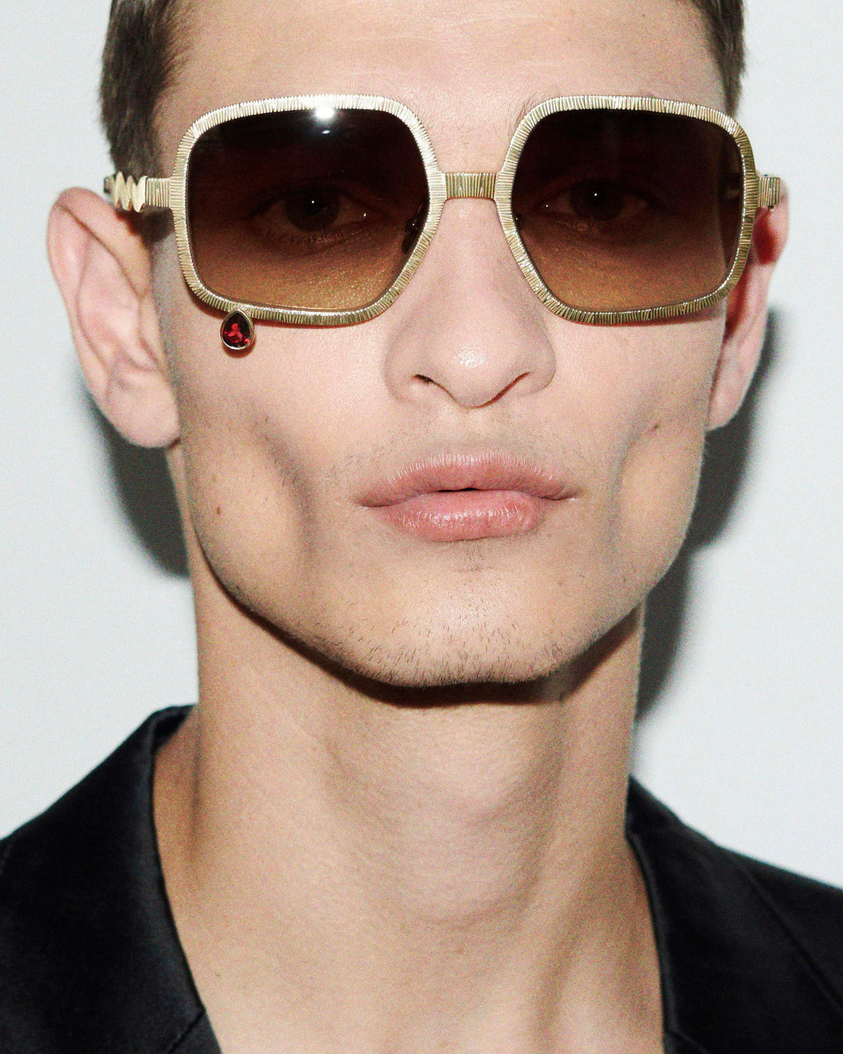 A close-up of a model wearing oversized square sunglasses with a textured gold frame and brown tinted lenses, paired with a black shirt against a plain light background.