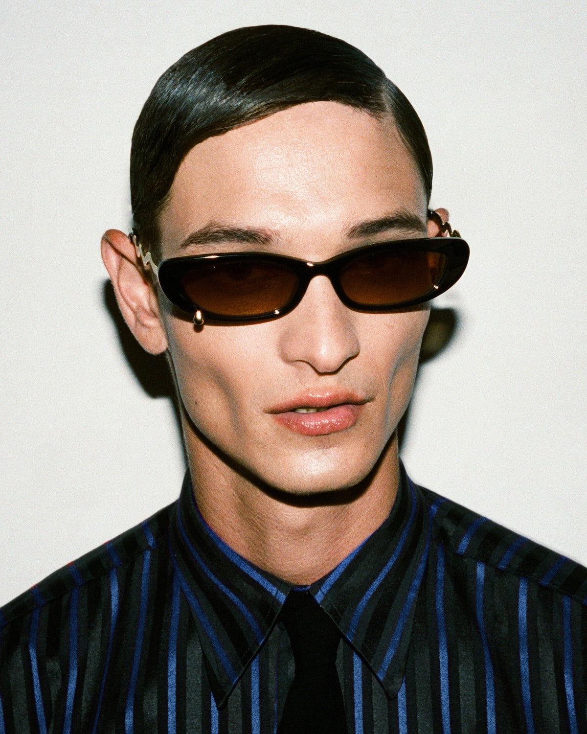 A close-up portrait of a model wearing dark brown cat-eye sunglasses with gold-tone metal frames and a blue-and-black striped shirt against a plain light background.