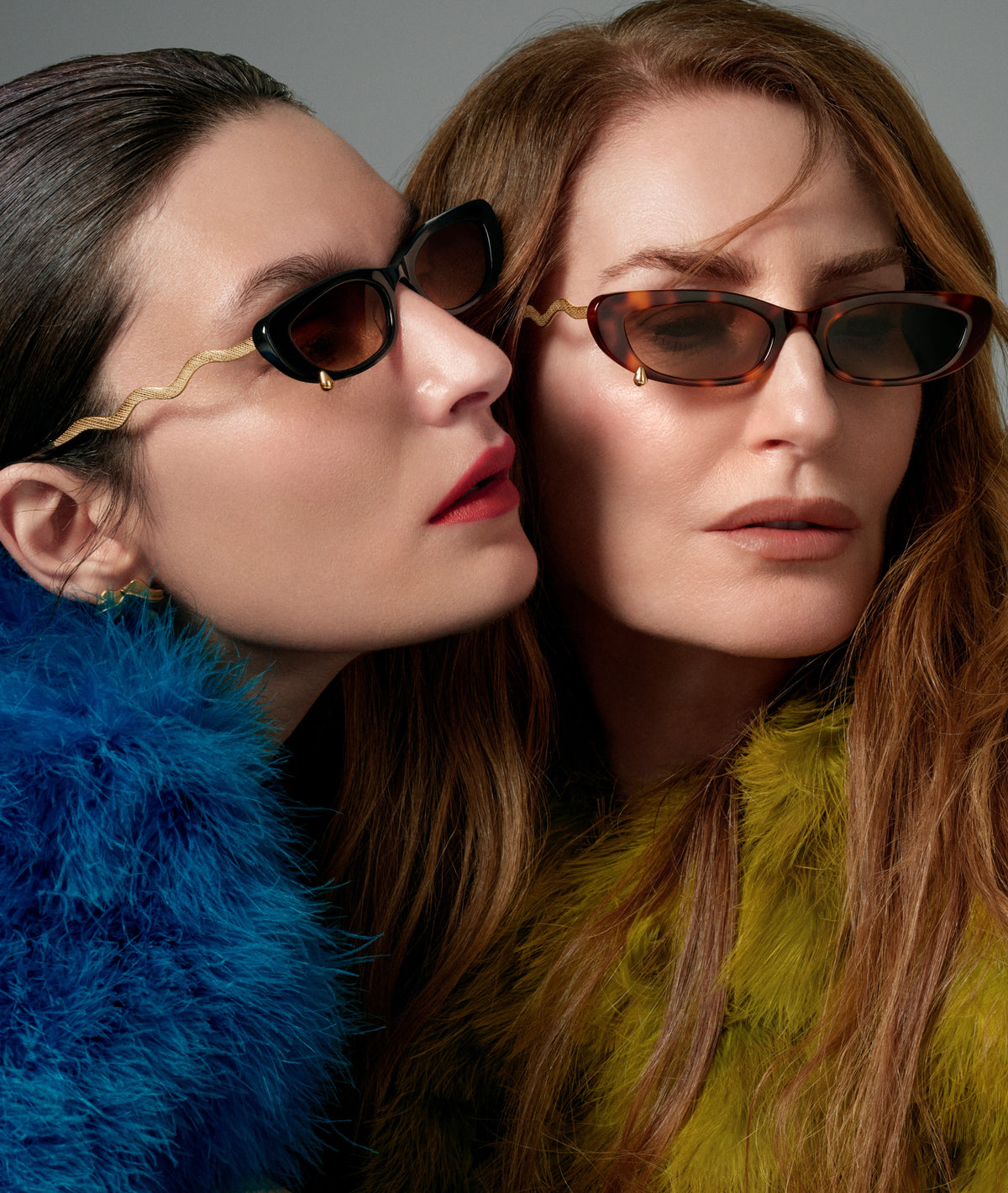 Two women wearing stylish cat-eye sunglasses with gold and tortoiseshell frames, styled with vibrant blue and yellow faux fur collars against a neutral gray background.