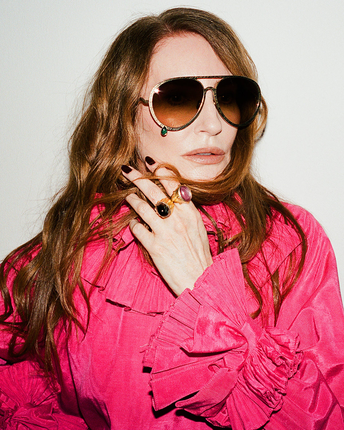A woman with long wavy light brown hair wearing aviator sunglasses with gold frames and brown lenses, paired with a bright pink ruffled blouse. She is posing with her hand near her face against a plain light background.