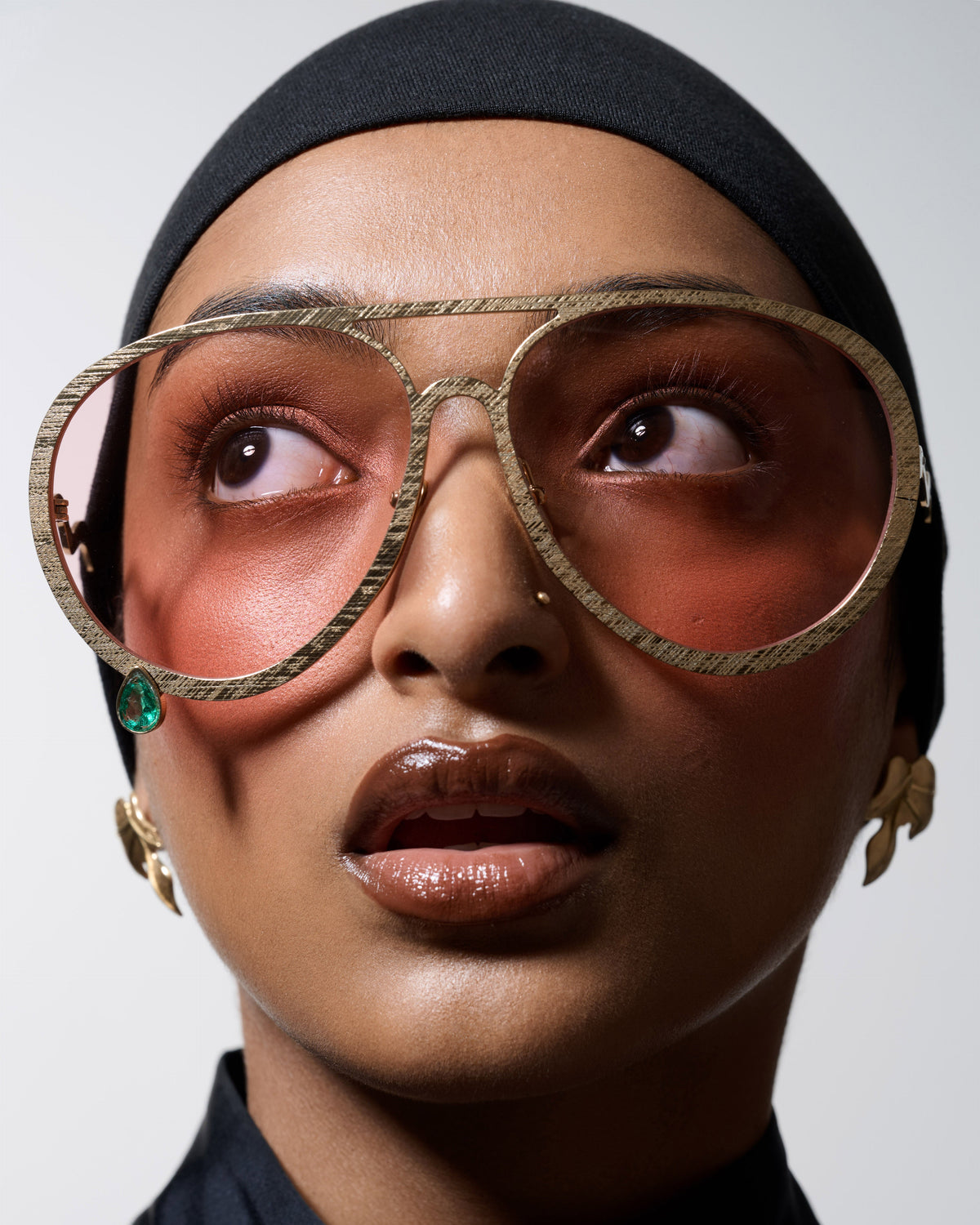 A close-up portrait of a model wearing oversized round aviator-style sunglasses with a textured gold-tone frame, peach-tinted lenses, and a small green jewel accent on the left side, against a light gray background.