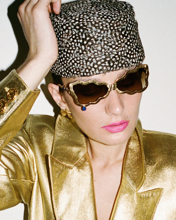 A close-up fashion shot of a model wearing a metallic gold satin blazer, a matching gold hat with white spots, and scalloped gold sunglasses against a plain light background.